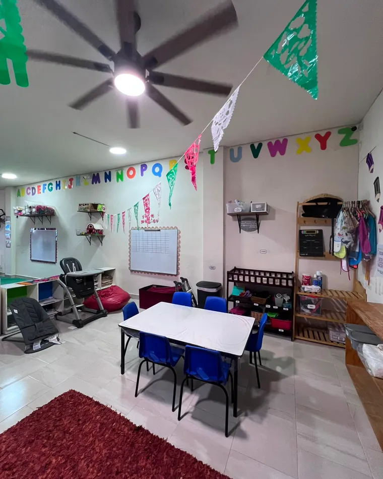 Otra vista del salón de kínder con mesas, estantes y abecedario.